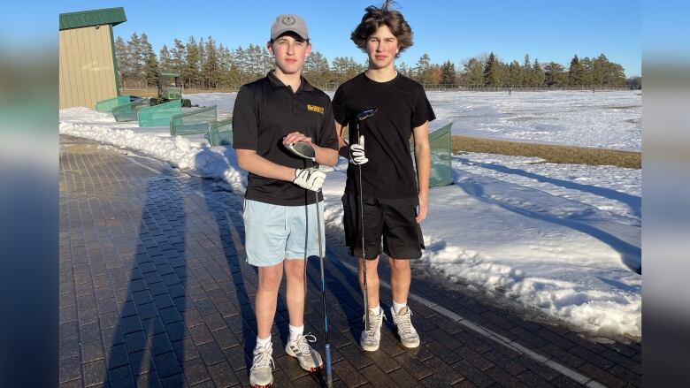 Two teenage boys stand side by side in shorts, holding golf clubs with snow in the background.