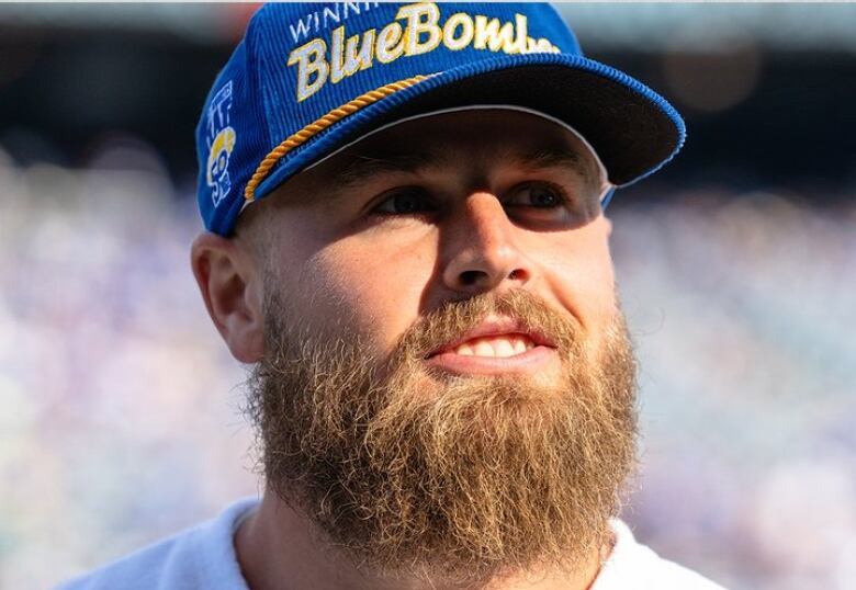 A man with a bushy beard and a blue baseball cap smiles and looks off to the side of the photo frame