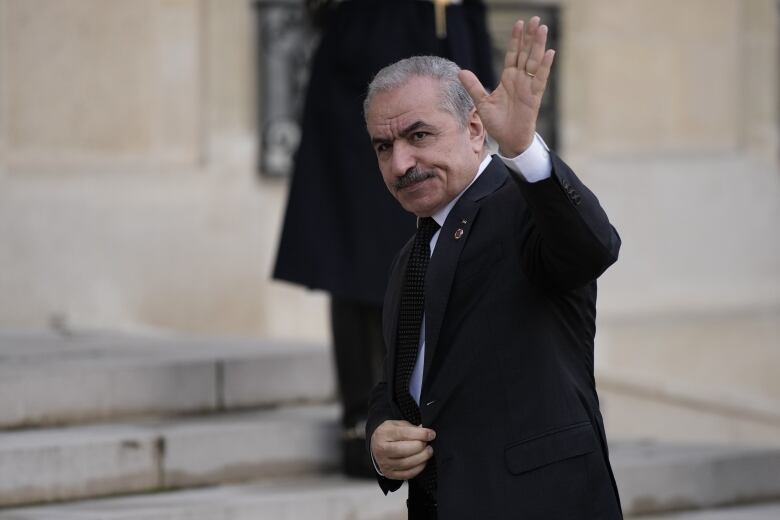 A man in a black suit waves as he walks toward a stone building.