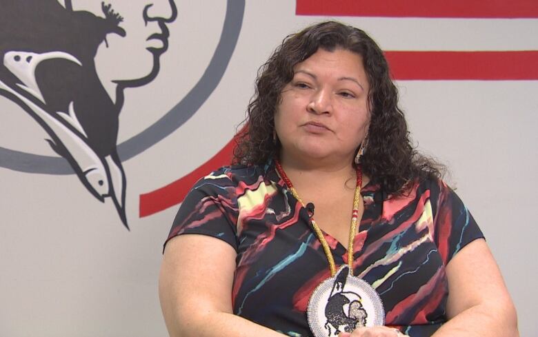 A First Nations woman wearing a multicolour t-shirt and a beaded medallion sits in front of a large logo on the wall.