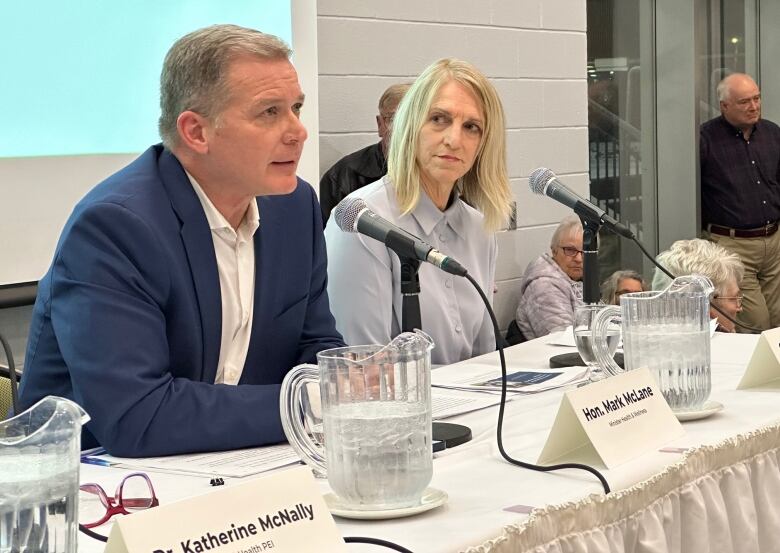 Man in blue business jacket speaks into a microphone as a woman with long blond hair looks on from the next seat at the podium table. 