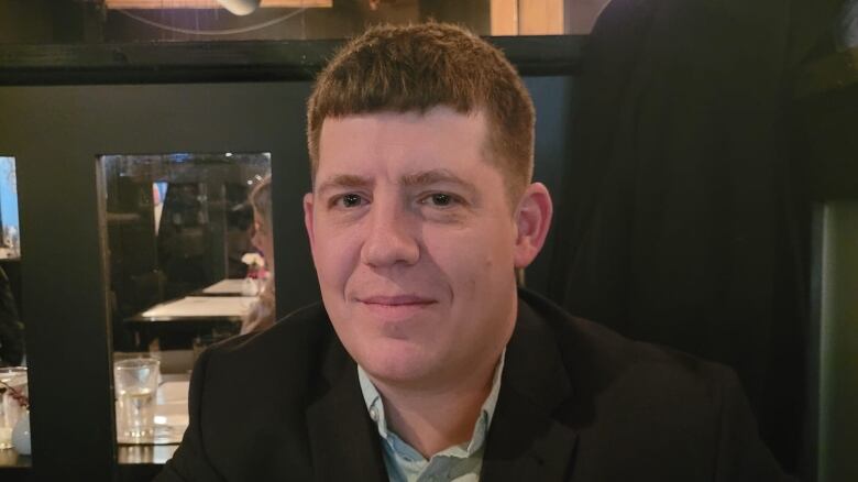 A man in about his 30s with short light brown hair and wearing a black sports coat and pale green button down shirt sits at a desk or table in a room with dark green walls that open to a higher ceiling with wooden rafters and pendant lighting. 