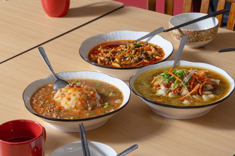 Three dishes sit on a table. Each different, they have varied orangey broth bases and are topped with meat, fish, and chopped up vegetables.