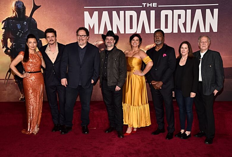 A long of well-dressed people stand on a red carpet. Behind them is a photowall bearing the words 