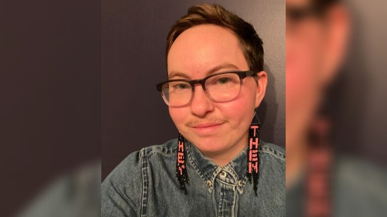 A person takes a selfie against a dark wall. They have short brown hair and glasses. They wear a denim jacket and they/them earrings.