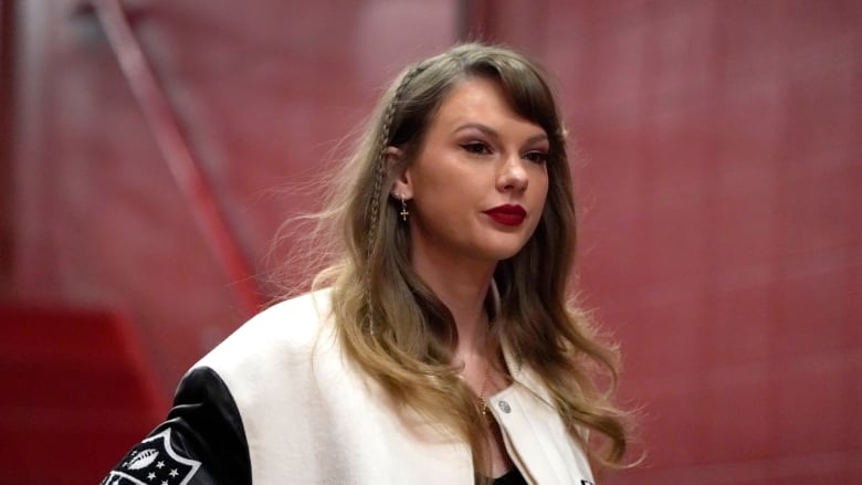 A blonde woman wearing a varsity jacket walks in a red corridor.