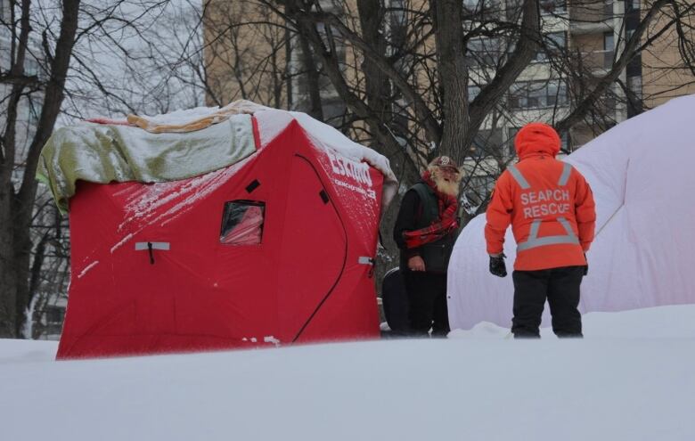 A man in a bright orange search and rescue jacket speaks to a homeless man outside his tent in a snowy city park.