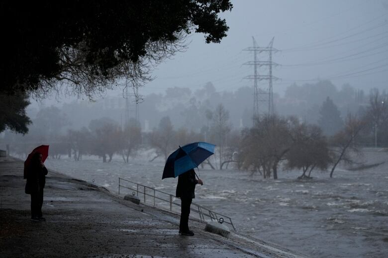 People with umbrellas look on as a storm causes a body of water to flow rapidly.