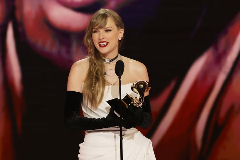 A blonde woman wearing a white dress with black gloves holds an award while speaking into a microphone.