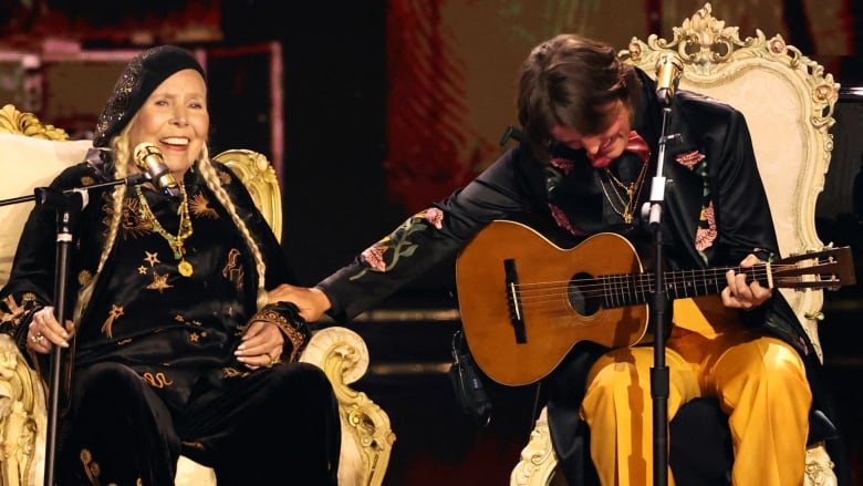 A woman with a guitar smiles as she bows her head and puts her hand on the forearm of another smiling woman. They both sit before microphones.