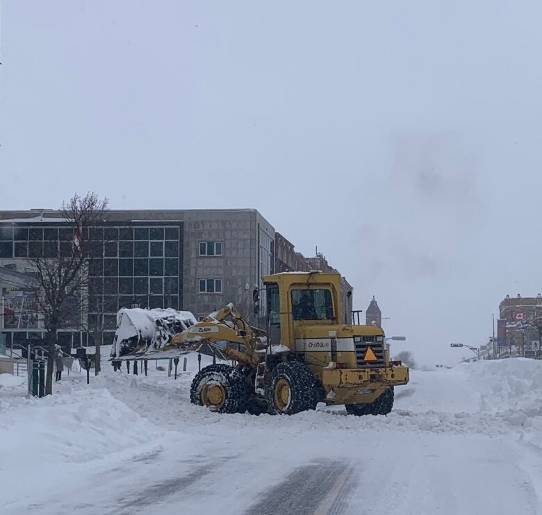 Tractor picks up snow on quiet street with buildings in background