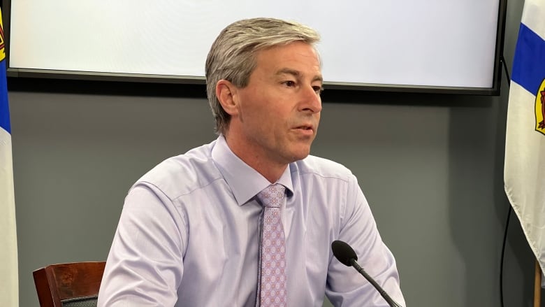 Man with a light grey shirt and mauve coloured tie sits at a desk with a microphone and glass of water in front of him.