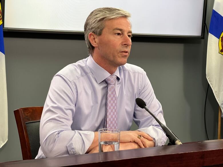 Man with a light grey shirt and mauve coloured tie sits at a desk with a microphone and glass of water in front of him.