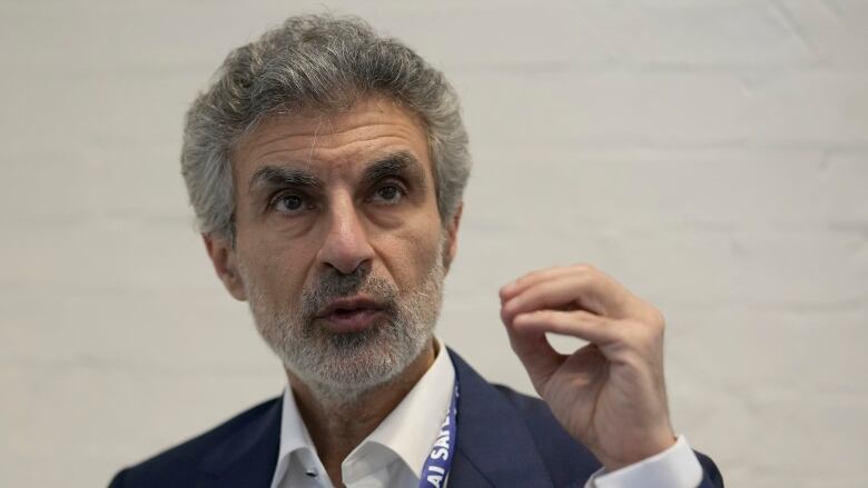 A man in a dark blazer and white shirt gestures with his hand while speaking.