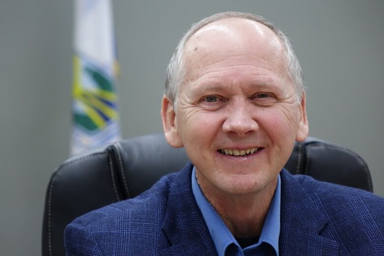 A balding man wearing a blue suit smiles while posing with a flag in the background.