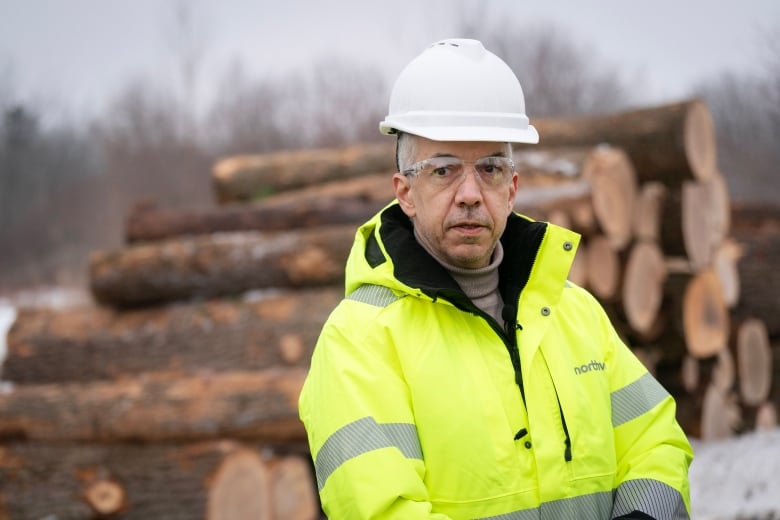 Man in safety glasses and a neon bright safety coat and white hard hat stands in front of a stack of felled trees.