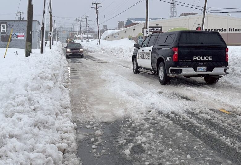 A black-and-white sport utility vehicle with police on the door drives down an icy street with large snowbanks on either side.