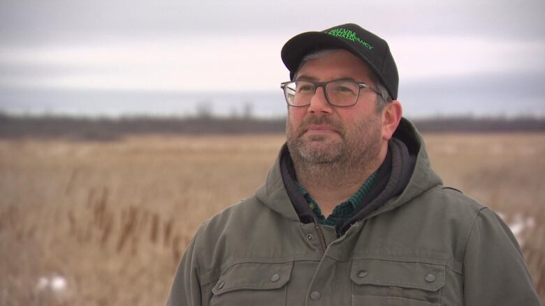 A man stand outdoors in front of a rural wetland property wearing a tan jacket with glasses.