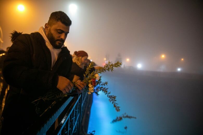 People gather along the railing of a bridge holding flowers on a foggy evening. 