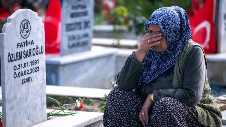 An older woman in a headscarf sits next to a grave, her face buried her hand. 