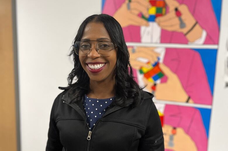 A woman with glasses stands in front of a mural depicting hands solving a rubics cube.