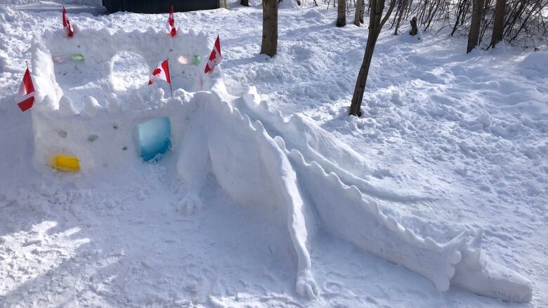 A dragon made of snow stretches across the ground, guarding a snow castle. Miniature Canadian flags stick up out of the castle.