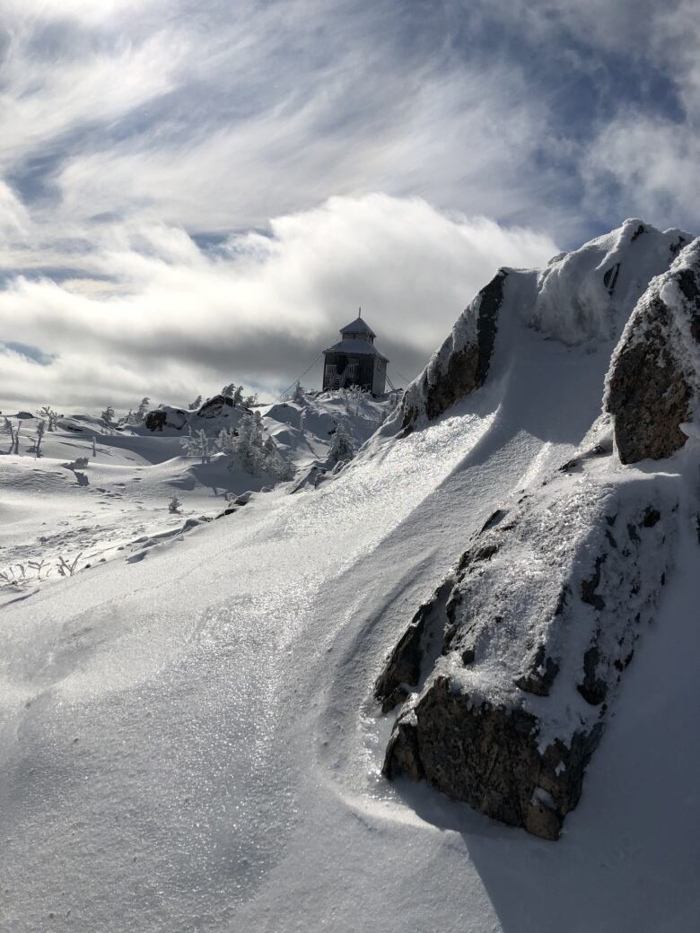 Snow and rock swirl together beneath a blue sky brushed over with clouds. In the distance is a small building.