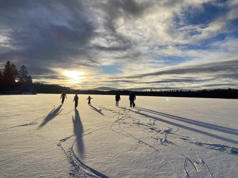 Five people skate over a lake dusted with snow as the sun sets in the background.