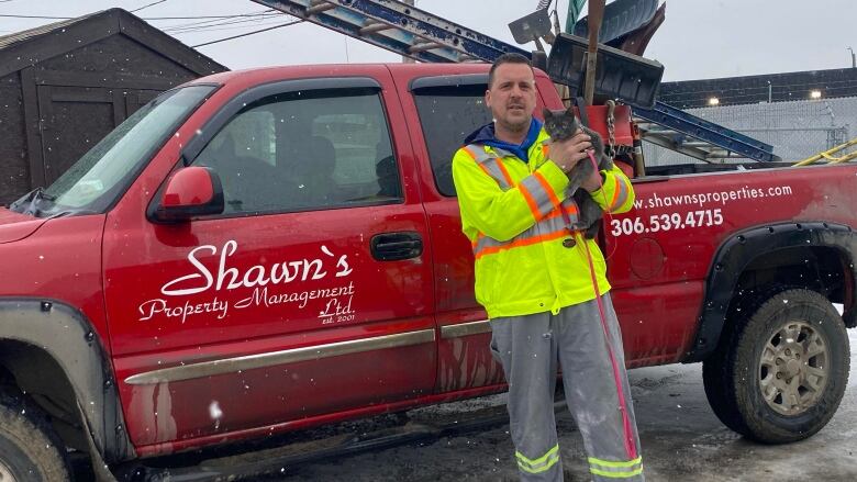 A man stands in front of a work truck smiling with a small adorable cat in his hands.