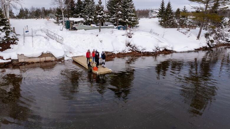 A group of people in winter jackets stands on a dock watching someone in the water with a rake, tonging for oysters. 