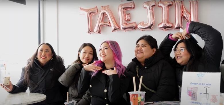 A woman with long pink hair, dressed in black, sits beside four other women, also dressed in black, and smiling broadly for the camera.