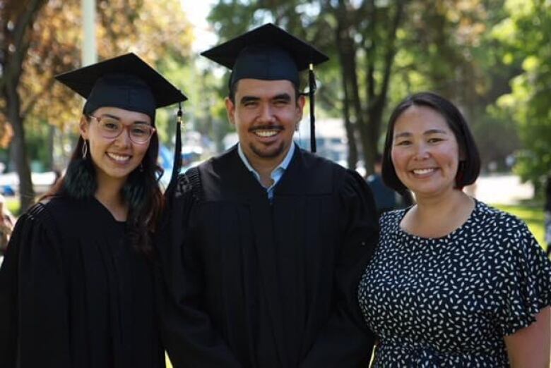 Three people smile at the camera. Two people wear graduation cap and gowns. 