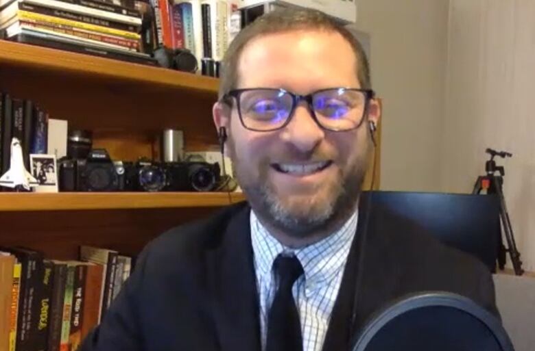 A man with a beard wearing a suit and tie sits in his office smiling.