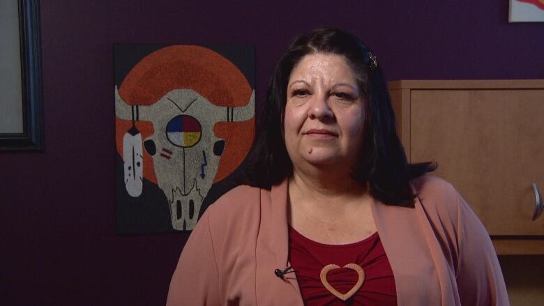 A woman with medium-length dark hair stands in front of art painted on the wall behind her. The painting features a bison skull, with a medicine wheel painted in the centre.