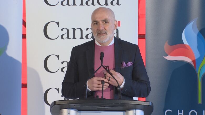 A man holding a pair of glasses stands at a podium speaking into a microphone. 