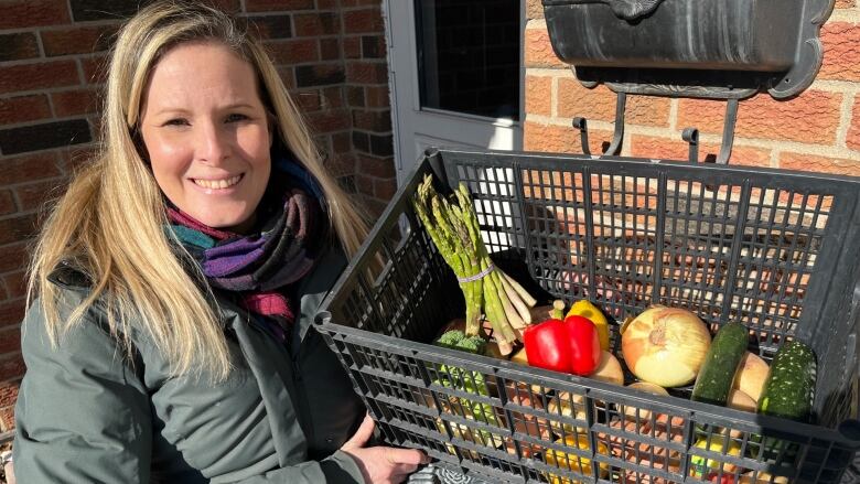 A woman stands in front of her house holding a box of vegetables and fruits. 