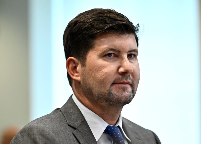 A man in a grey suit sits in front of a window.