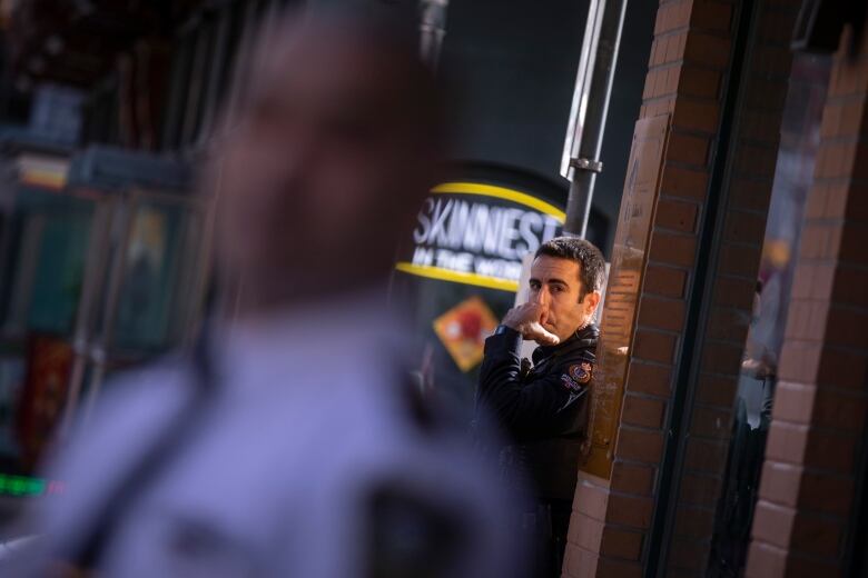 A police officer leans against a brick wall, with another pictured in silhouette.