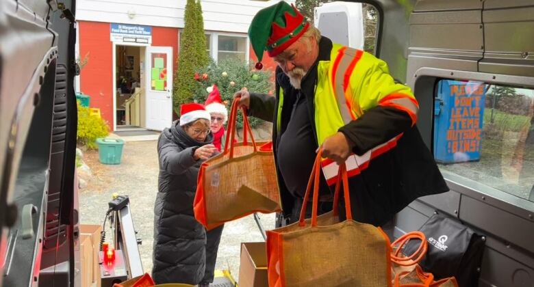 A white man with a beard wears a green elf hat, standing in the back end of a large van. He is carrying two brown bags full of food as more are placed around him