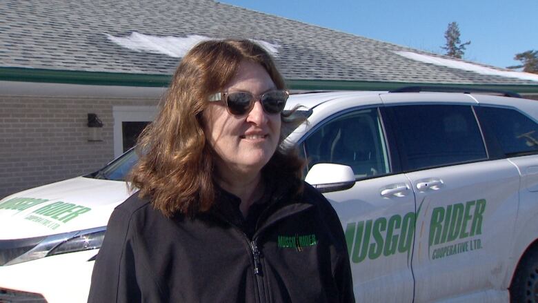 A white woman with shoulder-length brown hair and sunglasses stands outside in front of a white van with the MusGo logo on it