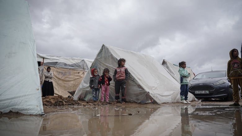 Children stand outside of tents in a muddy and wet camp ground.