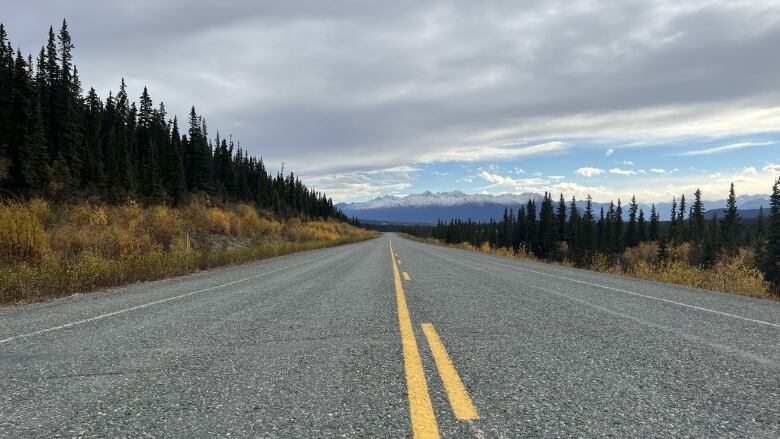 Looking down an empty highway with trees and mountains in the background.