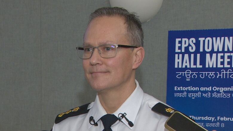 A man with glasses, wearing a police uniform, stands in front of a sign that says 