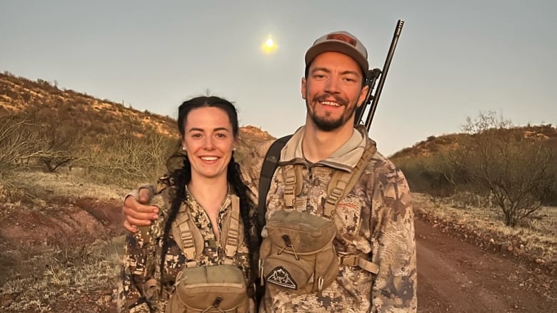 A couple both wearing camo stand smiling.