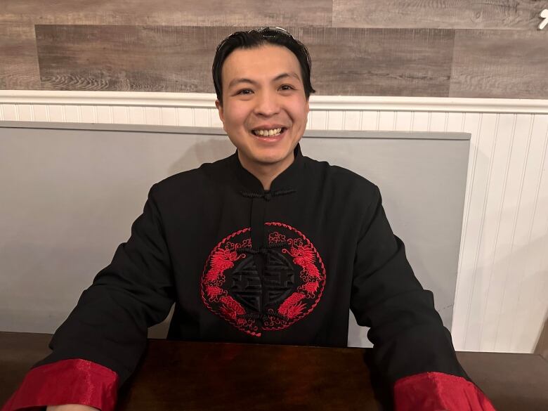 A man in a black and red long sleeve shirt sits smiling at the camera. 