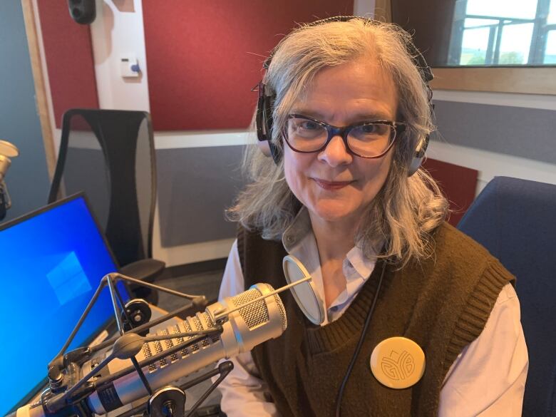 A woman with grey hair and glasses and wearing headphones, sits in front of a radio studio microphone. 