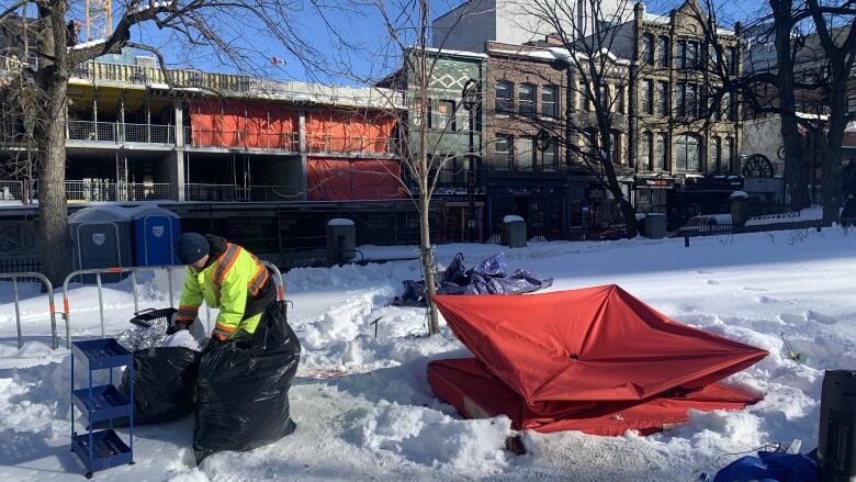 A man in neon safety clothing gathers two black garbage bags from the snow beside a deconstructed tent.