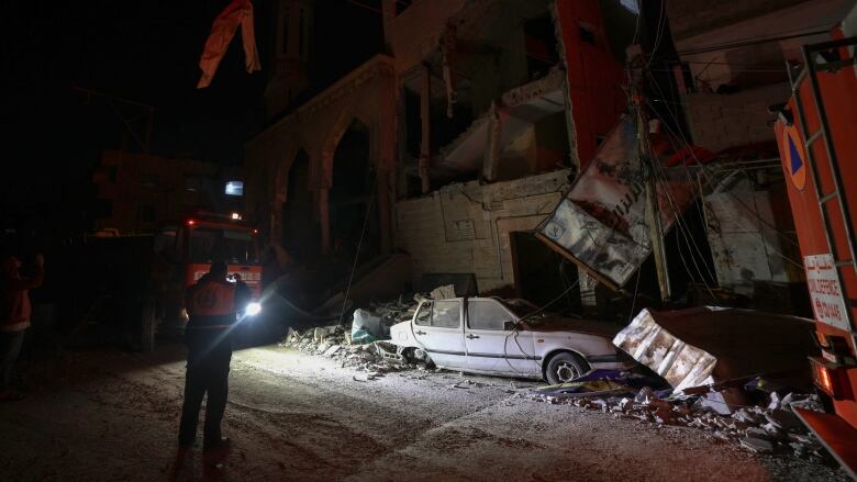 A worker shines a flashlight on a destroyed car covered in rubble. 