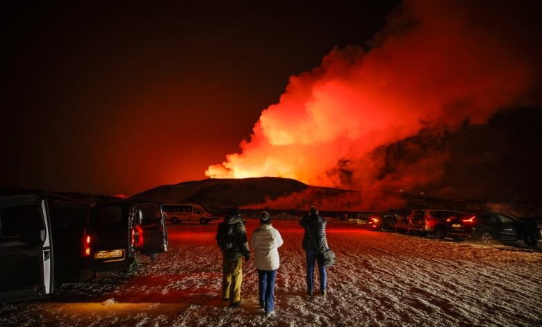 Three people stand in a parking lot as smoke and lava moves over the horizon.