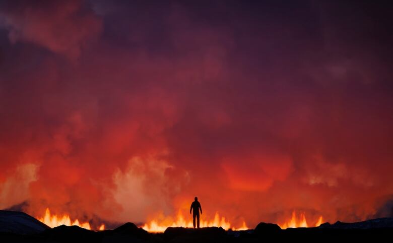 A person stands on the edge of an erupting volcano in Iceland as smoke and red flames fill the sky.